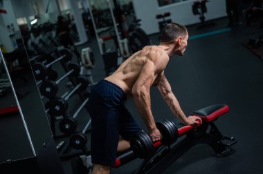 Gömleksiz bir adam spor salonunda dambıl sırasına dizer.