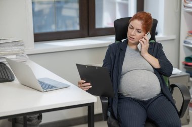 Ofiste cep telefonu kullanan ve kağıt tablet tutan hamile bir kadın.