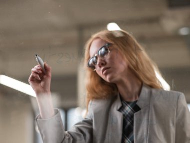 Gözlüklü beyaz kadın cam duvara İngilizce yazı yazıyor.