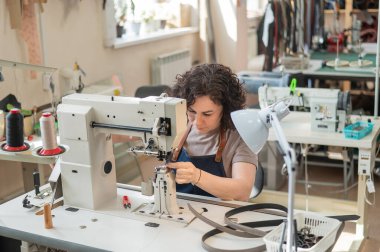 A woman tanner sews a leather belt on a sewing machine clipart