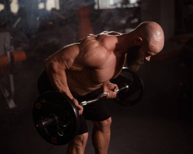 Beyaz, kel, üstsüz bir adam spor salonunda halterle egzersiz yapıyor.