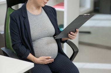 Ofisteki kağıt tablette belge okuyan hamile bir kadın. Karnına yakın çekim.