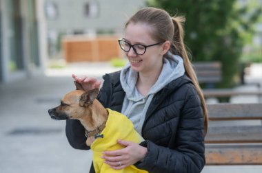 Genç beyaz bir kadın bir Rus oyuncak teriyeriyle yürüyor. Ceketli bir köpek sahibi bankta oturuyor.