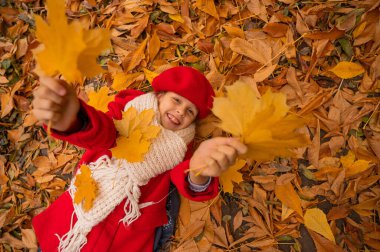 Kırmızı paltolu ve bereli beyaz bir kızın yukarıdan görünüşü sarı yaprakların üzerinde yatıyor. Sonbaharda parkta yürüyüş
