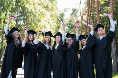 Mezuniyet elbisesi giymiş, açık havada diplomalarını gösteren bir sürü genç öğrenci.