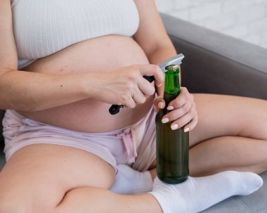 Kanepede oturan ve bira şişesini açan yüzü olmayan hamile kadın.