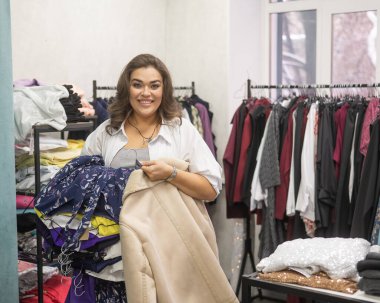 Büyük boy mağazada kucak dolusu kıyafet tutan mutlu şişman kadın.