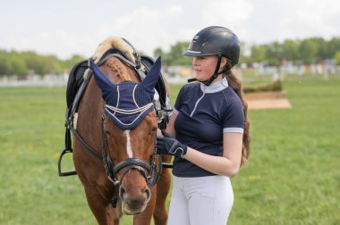 Genç bir kız bir binicilik yarışmasının önünde bir atın yanında duruyor.