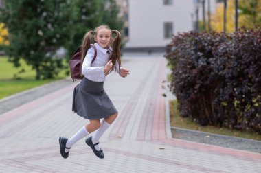 Üniformalı ve sırt çantalı mutlu beyaz kız okuldan sonra dışarı atlıyor.