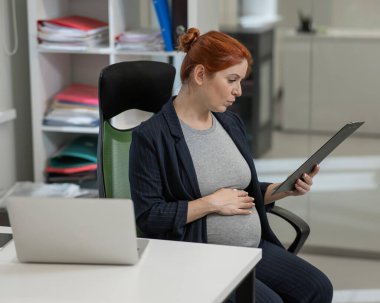 Ofisteki kağıt tablette belgeleri okuyan hamile bir kadın.