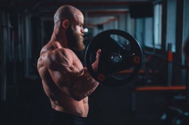 Beyaz, kel, üstsüz bir adam spor salonunda halterle egzersiz yapıyor. Ağırlıklarla bibik bukleler