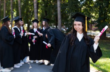 Mezuniyet elbisesi giymiş bir grup mutlu öğrenci dışarıda. Ön planda elinde diploması olan genç bir kız.