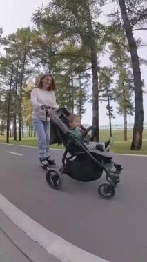 Beyaz kadın, yürümekte olan oğluyla bebek arabasında paten kayıyor. Dikey video