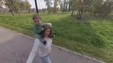 Beyaz bir kadın parkta yürürken oğlunu omuzlarında taşır.