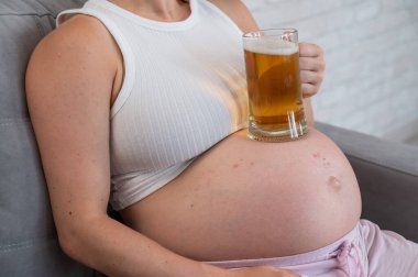 Yüzü olmayan hamile bir kadın kanepeye oturur ve karnına bir bardak bira koyar. Deri döküntüsü