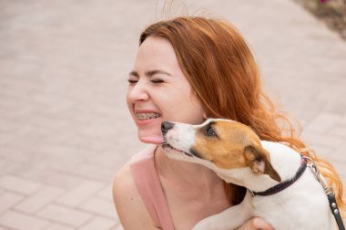 Köpek Jack Russell Terrier sahibinin yüzünü açık havada yalıyor. Dişlerinde diş teli olan kız.