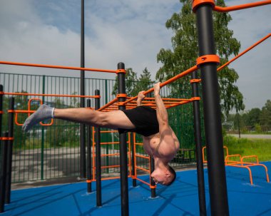 Üstsüz erkek sporcu sırt üstü uzanıyor. Açık havada yatay bara asılıyor.