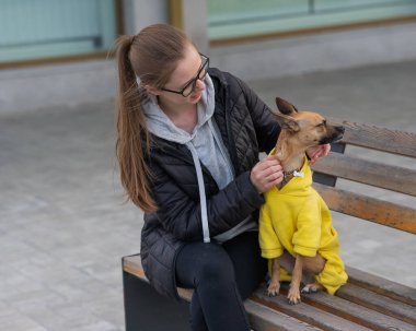 Genç beyaz bir kadın bir Rus oyuncak teriyeriyle yürüyor. Ceketli bir köpek sahibi bankta oturuyor.