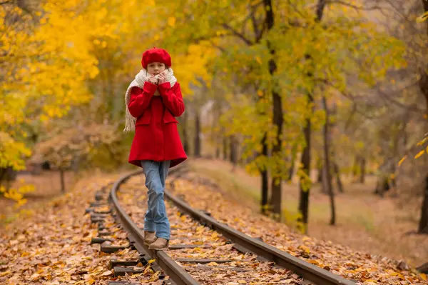 Kırmızılı ve bereli beyaz kız sonbaharda parkta tren yolu boyunca yürür.