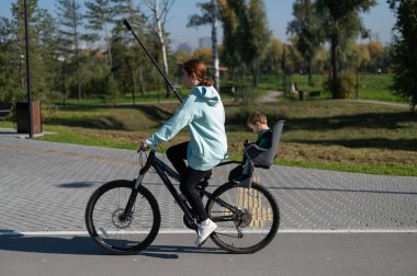 Beyaz kadın bisiklete biniyor. Yanında da oğlu var. Parkta çocuk koltuğunda oturuyor.