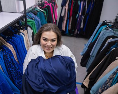 Şişko yanaklı bir kadının kucak dolusu giysiyi büyük boy bir mağazada tutuşu.