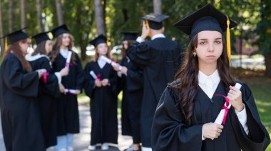 Mezuniyet elbisesi giymiş bir grup mutlu öğrenci dışarıda. Genç bir kızın elinde diploması var.