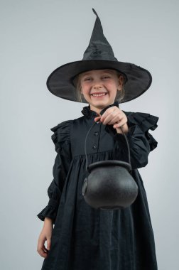 Beyaz arka planda cadı kostümü giymiş beyaz bir kızın elinde kazan tutan portresi. Dikey fotoğraf