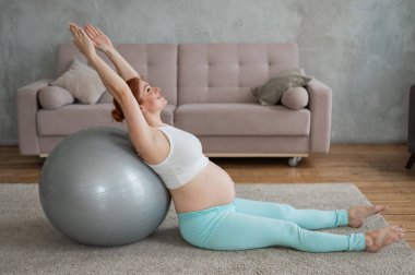 Hamile kızıl saçlı kadın evde fitness topu egzersizleri yapıyor.