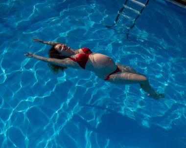 Havuzda kırmızı bikinili hamile bir kadın.