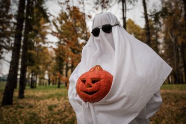 Woman in white sheet holding jack olantern against autumn forest background. Halloween costume clipart