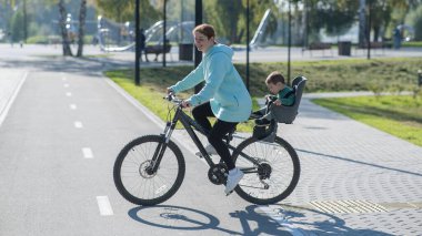 Beyaz kadın bisiklete biniyor. Yanında da oğlu var. Parkta çocuk koltuğunda oturuyor.