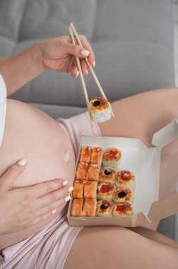 Hamile bir kadın kanepede oturur ve bir kutudan ekmek yer. Yemek servisi. Karnını kapat.