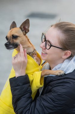 Genç beyaz bir kadının portresi ceketli küçük bir köpeğe sarılıyor. Dikey fotoğraf