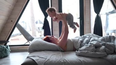 Kızıl saçlı beyaz kadın, panoramik pencereli bir kır evinde bebeğiyle yatakta yatıyor.