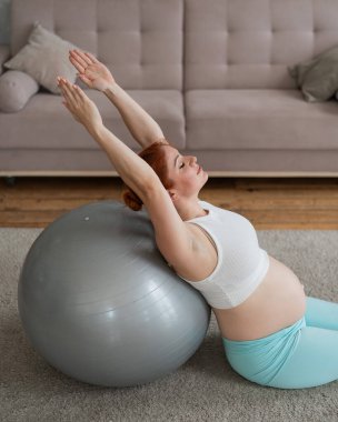 Hamile kızıl saçlı kadın evde fitness topu egzersizleri yapıyor.