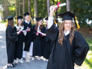 Mezuniyet elbisesi giymiş bir grup mutlu öğrenci dışarıda. Ön planda elinde diploması olan genç bir kız.