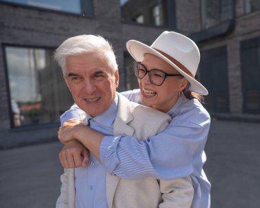 Şapkalı ve gözlüklü bir kadın beyaz ceketli yaşlı bir adama arkadan sarılıyor. Olgun insanların romantik ilişkileri