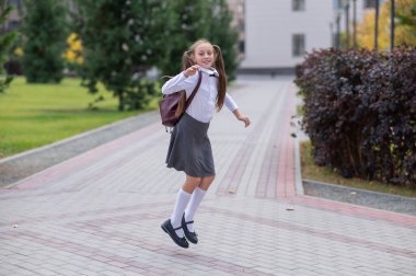 Üniformalı ve sırt çantalı mutlu beyaz kız okuldan sonra dışarı atlıyor.