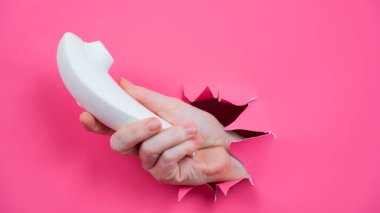 Kadın eli, beyaz vakum klitoral vibratörünü pembe kağıt arkaplanından dışarı çıkarıyor.