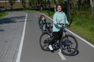 Küçük oğluyla bisiklete binen beyaz bir kadın arkasında çocuk koltuğunda oturuyor.