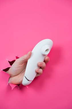 Kadın eli, beyaz vakum klitoral vibratörünü pembe kağıt arkaplanından dışarı çıkarıyor.