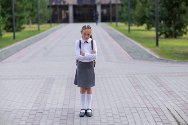 Üniformalı, sırt çantalı, kollarını göğsüne kavuşturmuş üzgün, beyaz bir kız okuldan sonra dışarıda duruyor.