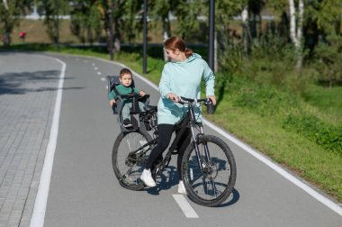 Küçük oğluyla bisiklete binen beyaz bir kadın arkasında çocuk koltuğunda oturuyor.