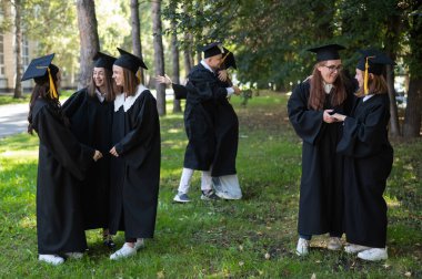 Mezuniyet elbisesi giymiş bir grup mutlu öğrenci dışarıda. Ön planda, iki genç kız birbirini tebrik ediyor ve sarılıyorlar.