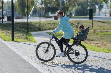 Küçük oğluyla bisiklete binen beyaz bir kadın arkasında çocuk koltuğunda oturuyor.