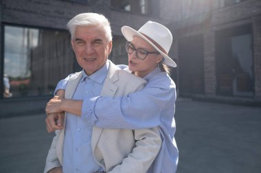 Şapkalı ve gözlüklü bir kadın beyaz ceketli yaşlı bir adama arkadan sarılıyor. Olgun insanların romantik ilişkileri