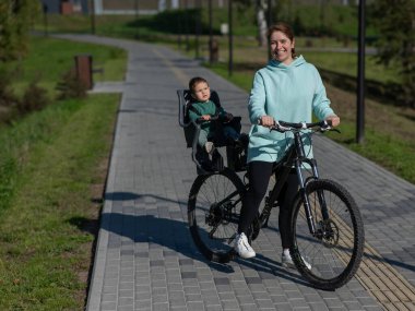 Küçük oğluyla bisiklete binen beyaz bir kadın arkasında çocuk koltuğunda oturuyor.