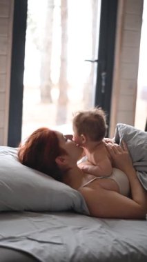 Kızıl saçlı beyaz kadın, panoramik pencereli bir kır evinde bebeğiyle yatakta yatıyor. Dikey video