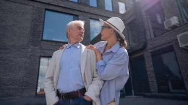 Şapkalı ve gözlüklü bir kadın beyaz ceketli yaşlı bir adama arkadan sarılıyor. Olgun insanların romantik ilişkileri