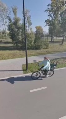 Beyaz bir kadın parkta yeni yürümeye başlayan oğluyla bisiklet sürüyor. Dikey video